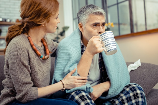 温かい飲み物。ソファの上で彼の妻の近くに座っている間熱いお茶を飲む素敵な病人