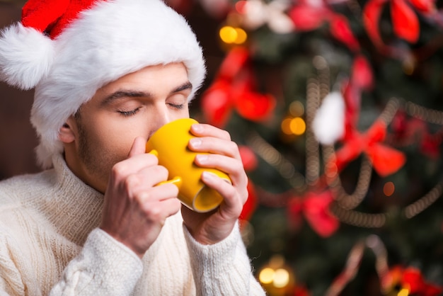 冬の夜の温かい飲み物。バックグラウンドでクリスマスツリーとホットドリンクを飲むサンタ帽子のハンサムな若い男