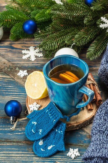 Hot drink with crocheted funny mittens. Traditional winter decor, fir tree branches, snowflakes. Festive seasonal arrangement in blue tones, old wooden background, close up