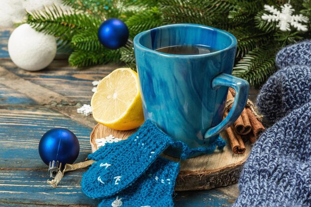 Hot drink with crocheted funny mittens. Traditional winter decor, fir tree branches, snowflakes. Festive seasonal arrangement in blue tones, old wooden background, close up