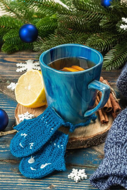 Photo hot drink with crocheted funny mittens. traditional winter decor, fir tree branches, snowflakes. festive seasonal arrangement in blue tones, old wooden background, close up