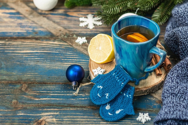 Hot drink with crocheted funny mittens. Traditional winter decor, fir tree branches, snowflakes. Festive seasonal arrangement in blue tones, old wooden background, copy space