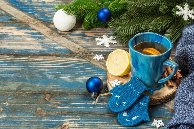 Hot drink with crocheted funny mittens. Traditional winter decor, fir tree branches, snowflakes. Festive seasonal arrangement in blue tones, old wooden background, copy space