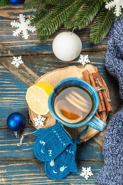 Hot drink with crocheted funny mittens. Traditional winter decor, fir tree branches, snowflakes. Festive seasonal arrangement in blue tones, old wooden background, top view