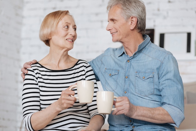 Hot feelings. Two elderly charismatic people spending some time together while sitting on bed and chatting with cups in their hands