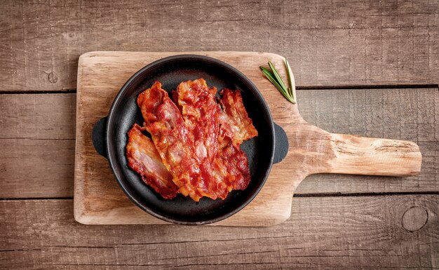 Hot fried crispy bacon slices and rosemary sprig in skillet on wooden