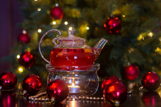 Hot fruit drink in a transparent teapot with a candle on the background of New Year's decoration.