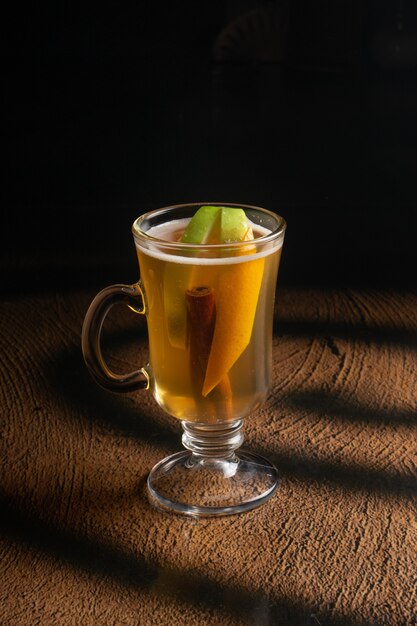 Hot fruit tea with apple slices, orange peel and a stick of cinnamon in a clear glass glass with a handle.