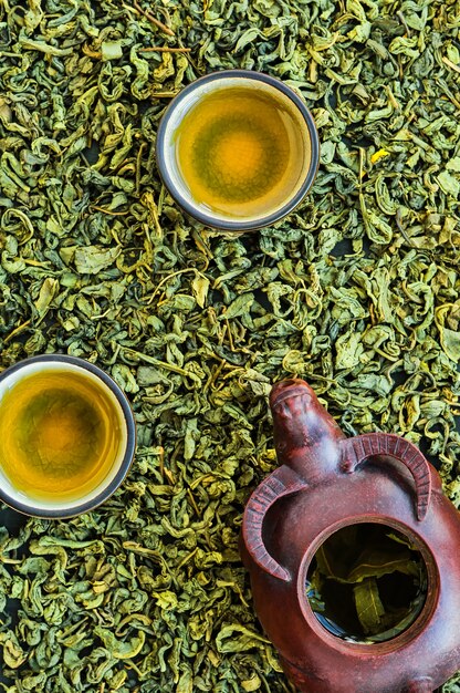 Горячий зеленый чай в чашках, керамический чайник на каменном столе, сервировка. На столе рассыпаны перебродившие чайные листья. Вид сверху. Минимальный