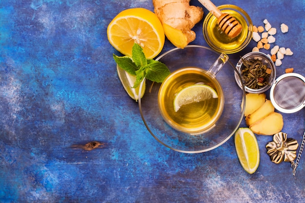 Hot green tea in a glass cup served with lemon, ginger, lime, honey and mint on grunge blue wooden background. Top view.