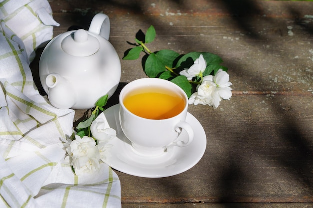 Hot green tea in a teapot and cup with a branch of jasmine flowers blossom  and white towel on rough rustic brown wooden background.  Nature healthy slow life concept. Copy space, top view.