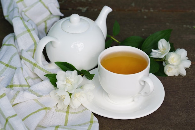 Hot green tea in a teapot and cup with a branch of jasmine flowers blossom  and white towel on rough rustic brown wooden background.  Nature healthy slow life concept.