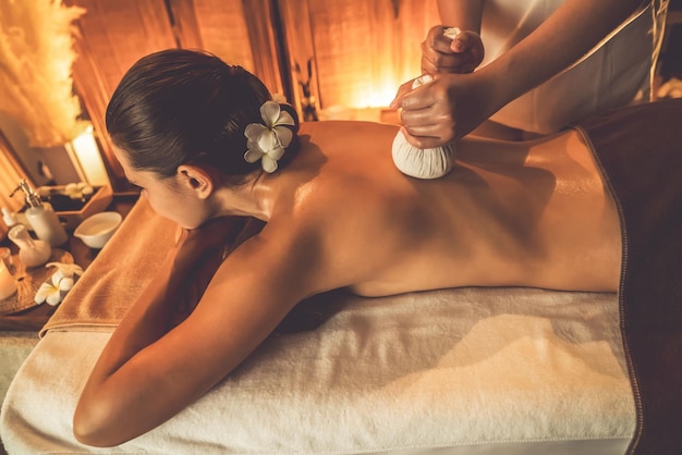 Hot herbal ball spa massage body treatment masseur gently compresses herb bag on woman body Tranquil and serenity of aromatherapy recreation in warm lighting of candles at spa salon Quiescent