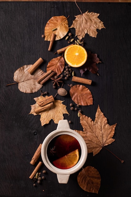 검은 나무 배경 위에 향신료, 오렌지, 가을 잎을 넣은 세라믹 냄비에 뜨거운 mulled 와인. 플랫 레이. 아늑한 따뜻한 음료