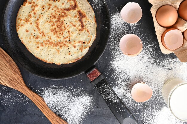 Горячий блин в черной сковороде на черном столе с мукой, молоком и яйцами.