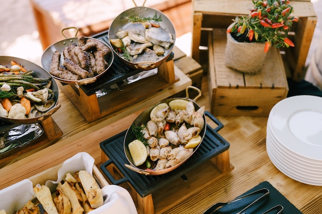 Hot pans with cooked food on a castiron stove on a wooden table with a bush of bitter pepper