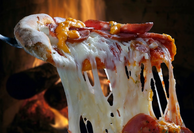 Hot pizza slice with melting cheese with wood oven in background.