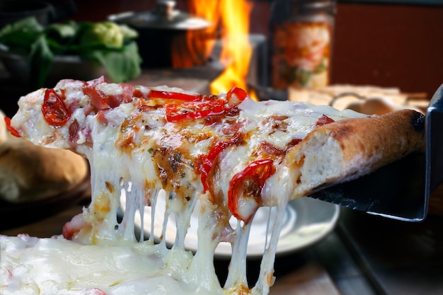 Hot pizza slice with melting cheese with wood oven in background.