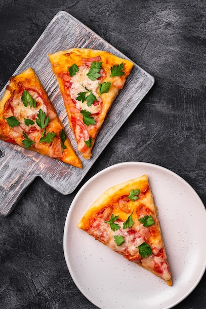 Hot pizza slices with mozzarella cheese, ham, tomato and parsley on wooden cutting board and plate, stone concrete table, top view