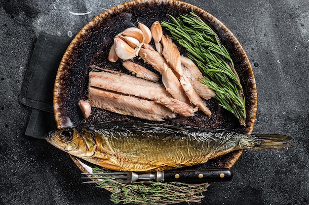 Hot Smoked herring fish fillet on rustic plate with thyme and rosemary. Black background. Top view.