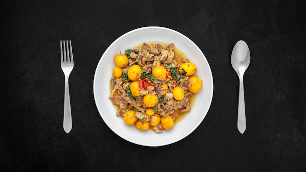 Photo hot and spicy stir fried chicken ovary egg with chicken entrails offal pluck variety meats organ meats and holy basil in white plate with fork and spoon on dark tone texture background top view