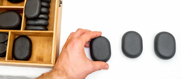 Hot stones for massage in a wooden box next to stone in the hand of masseur isolated on white background top view.