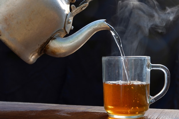 Чашка горячего чая имеет горячий пар с чайником. Помещен на старый деревянный стол, черный фон