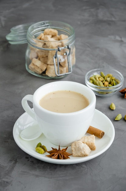 Photo hot tea with milk, cinnamon, cardamom, anise and other spices, indian masala tea in a white cup on a dark background. vertical, copy space.