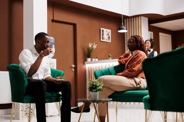 Hotel guests sitting in lounge area at modern resort, waiting to check in at reception desk. African american couple having accommodation in hotel lobby, employees providing excellent service.