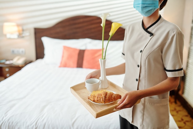 Hotel maid wearing medical mask when bringing tray with breakfast and flowers in hotel room