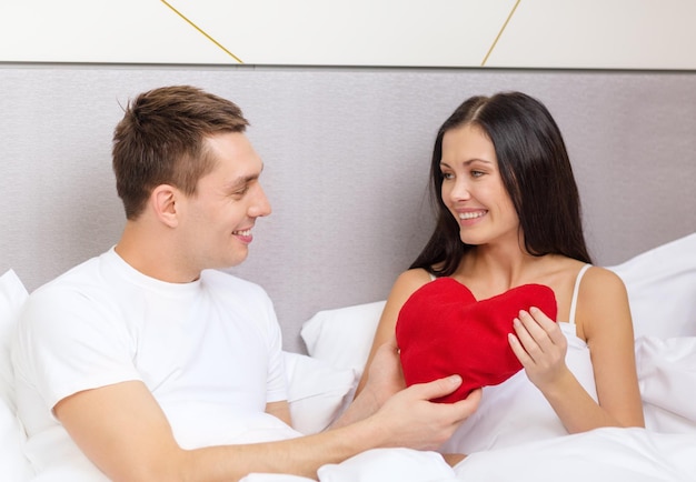 ホテル、旅行、関係、休日、幸福の概念-赤いハート型の枕とベッドで笑顔のカップル