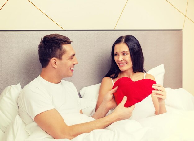 hotel, travel, relationships, holidays and happiness concept - smiling couple in bed with red heart-shaped pillow