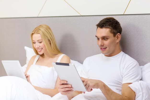 hotel, travel, relationships, technology, intermet and happiness concept - smiling couple in bed with tablet computers