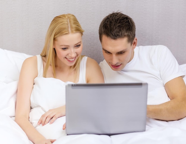 hotel, travel, relationships, technology, internet and happiness concept - smiling couple in bed with laptop