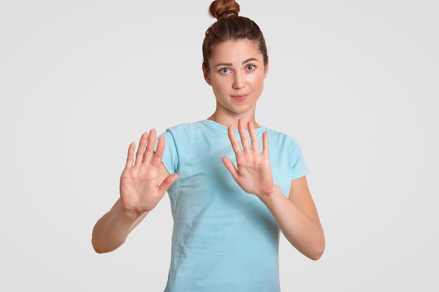 Hou op! De vrij jonge vrouw met aantrekkelijke blik, houdt palmen over borst, maakt weigeringsgebaar, gekleed in toevallige t-shirt, die over wit wordt geïsoleerd. Mensen, jeugd en lichaamstaal concept