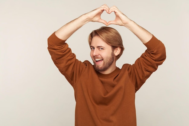 Hou van jou. Portret van een gelukkige knappe man met baard die een sweatshirt draagt met een gebaar in de vorm van een hart