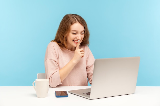 Houd het alsjeblieft geheim. Positieve jonge vrouw in vrijetijdskleding sussend met stiltegebaar, pratend op videogesprek, online communicatie via laptop op kantoor aan huis. binnen studio-opname geïsoleerd