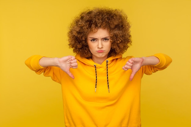 Houd niet van portret van ontevreden gefrustreerde krullendharige vrouw in stedelijke stijl hoodie met duimen naar beneden kritiek ontevreden met vreselijke service indoor studio opname geïsoleerd op gele achtergrond