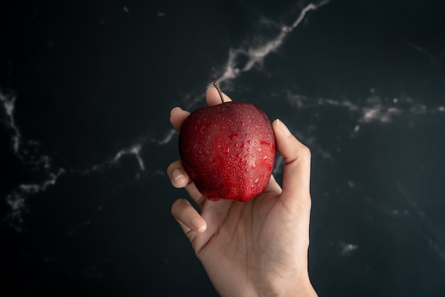 Foto houdend verse sappige rode appel met waterneveldruppeltjes op appel in hand over een zwart marmeren oppervlakte. bovenaanzicht plat lag samenstelling.