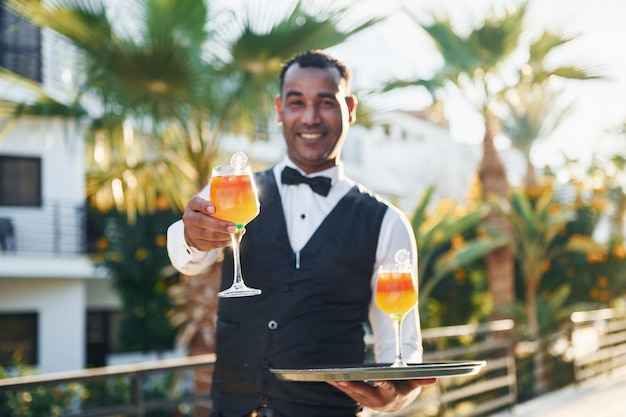 Houdt cocktails. Zwarte ober in formele kleding is op zonnige dag buiten aan het werk