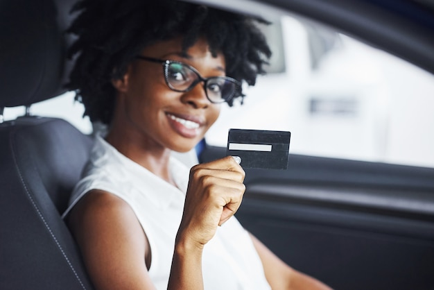 Houdt creditcard. Jonge Afro-Amerikaanse vrouw zit binnenkant van nieuwe moderne auto.