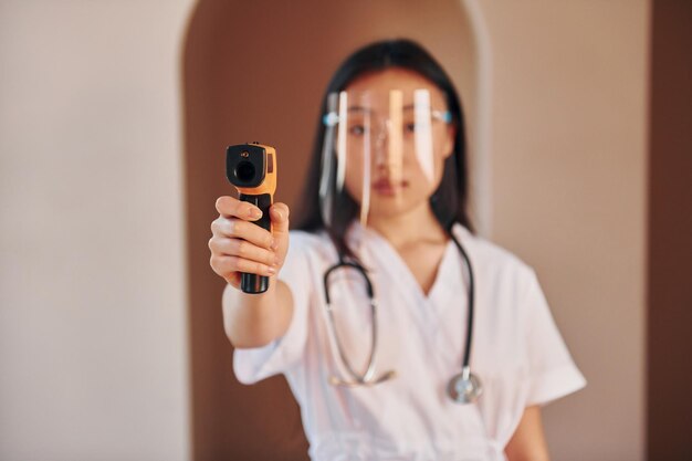 Houdt een infraroodthermometer vast Jonge serieuze aziatische vrouw die binnenshuis staat