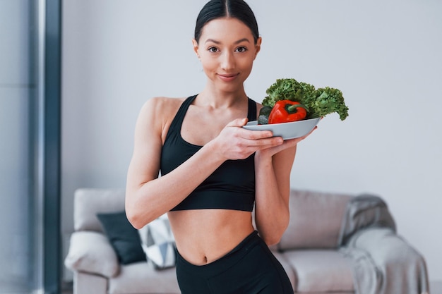 Houdt gezond voedsel in handen Jonge vrouw met slanke lichaamsvorm in zwarte sportkleding heeft thuis een fitnessdag binnenshuis