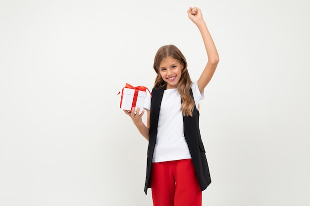 Houdt het tiener Kaukasische meisje in zwart jasje, wit overhemd en rode broek een witte doos met gift en verheugt zich geïsoleerd op wit