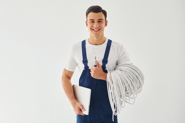 Houdt kabels notitieblok en schroevendraaier Mannelijke werknemer in blauw uniform staande binnenkant van studio tegen witte achtergrond