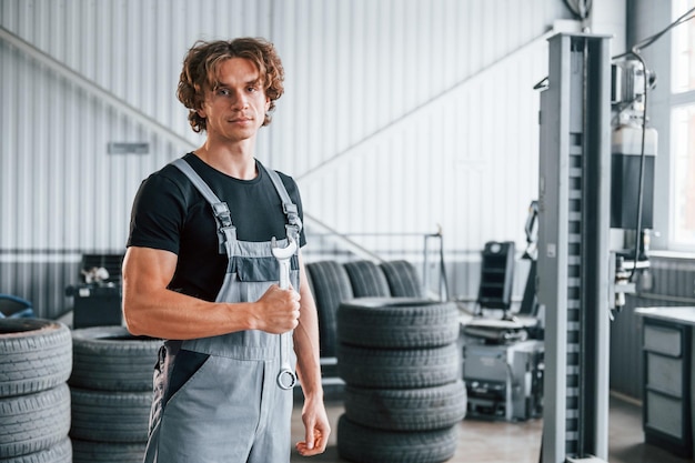 Houdt moersleutel in de hand volwassen man in grijs gekleurd uniform werkt in de autosalon