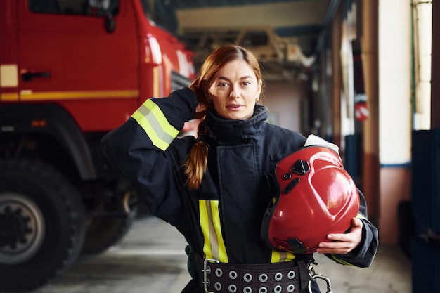 Houdt rode hoed in handen Vrouwelijke brandweerman in beschermend uniform staat in de buurt van vrachtwagen