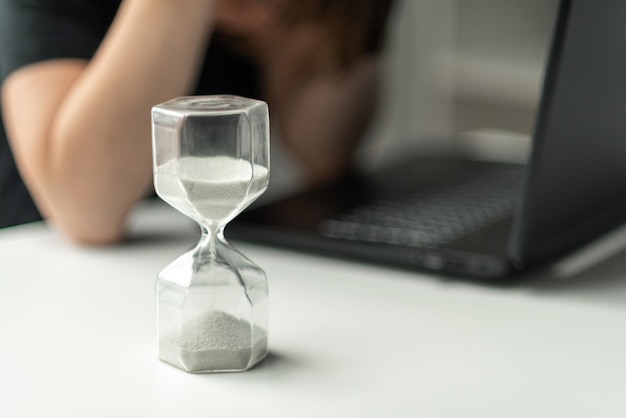 Photo hourglass in the foreground a woman is sitting in front of a laptop and is stressed