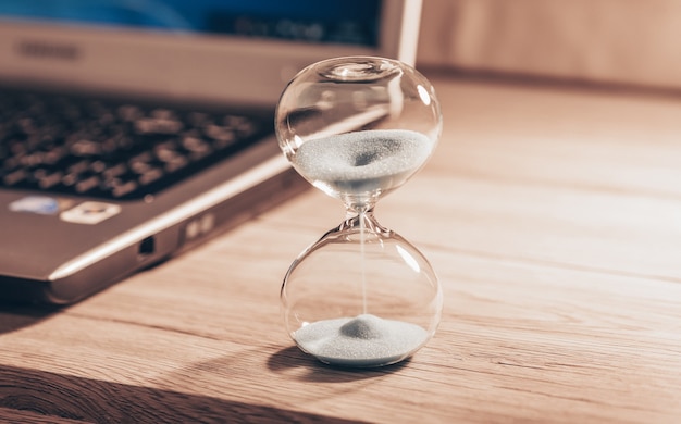 Photo hourglass on a light wooden table with a laptop or computer included. time slips through my fingers.