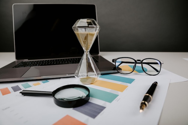 Фото Песочные часы на рабочем столе с диаграммной бумагой и ноутбуком бизнес-концепция